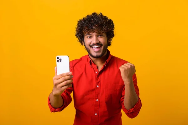 Happy satisfied man holding smartphone and smiling making yes gesture, celebrating online lottery or giveaway victory. Indoor studio shot isolated on orange — Fotografia de Stock