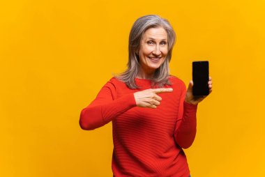 Sarı arka planda tek başına duran gri saçlı kadın reklam için akıllı telefonu işaret ediyor. Model resmi