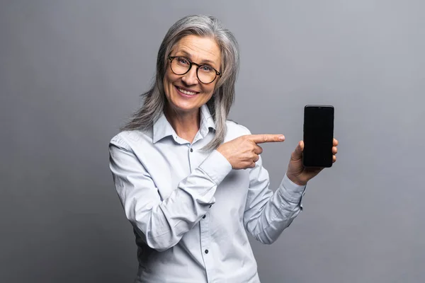 Mature gray-haired businesswoman wearing formal clothes showing smartphone and pointing with a finger on the screen with blank space for advertisement, standing isolated — ストック写真