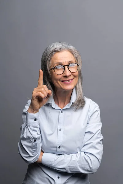 Vertical shot of the smiling mature gray-haired female business woman isolated on grey background points finger up. Lady paying attention — ストック写真