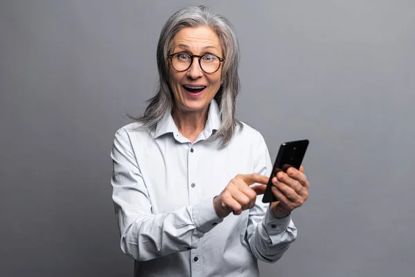 Здивована ділова жінка в офіційній сорочці, вказуючи пальцем на смартфон, відчуваючи себе шоковано новими функціями телефону. Внутрішній студійний знімок ізольовано на сірому — стокове фото