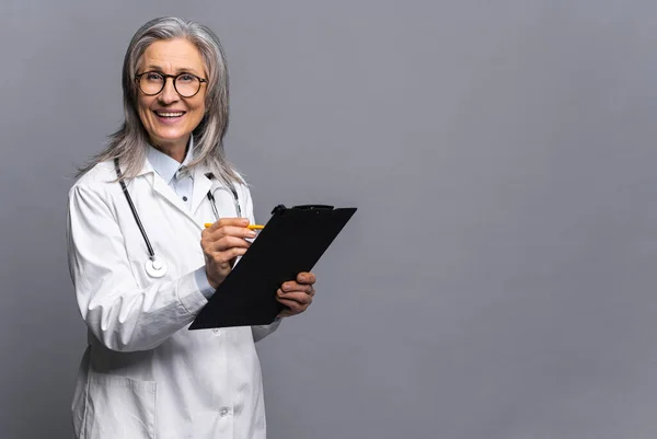 Senior doctor in white medical gown with stethoscope on shoulders taking notes standing with clipboard isolated on gray. Mature physician therapist writing down treatment plan — Foto Stock