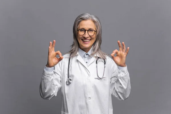 Cheerful senior mature female doctor with a stethoscope in a medical gown posing isolated over gray background, showing ok gestures. Smiling and welcoming healthcare worker — Stock Photo, Image