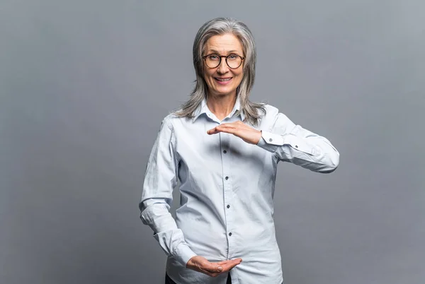 Successful senior mature gray-haired businesswoman gesturing with hands showing big and large size sign, measure symbol isolated on gray. Elder female in glasses standing — ストック写真