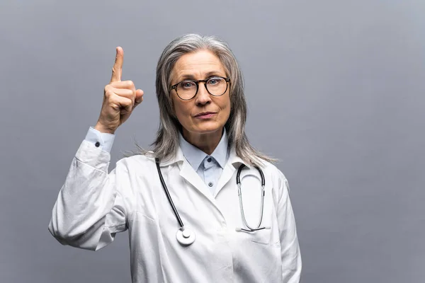 Smiling mature gray-haired female doctor in white medical gown, with stethoscope isolated on grey background points finger up. MD pays attention at health — Fotografia de Stock