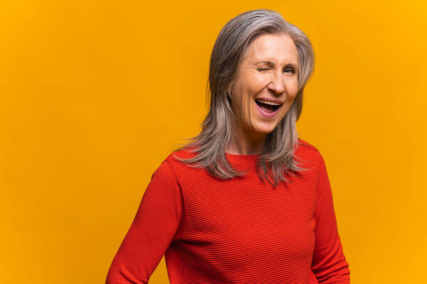 Charming senior woman laughing toothy. Elegant mature modern gray-haired lady in red jumper standing and blinking eye as having cunning idea, winking and flirting to camera. Happy lifestyle concept