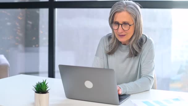Возбужденная седовласая пожилая женщина смотрит на экран ноутбука, сидя за столом в офисе — стоковое видео