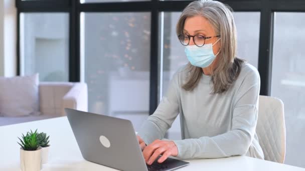 Γκρίζα μαλλιά ώριμη επιχειρηματίας φορώντας προστατευτική ιατρική μάσκα χρησιμοποιώντας φορητό υπολογιστή — Αρχείο Βίντεο