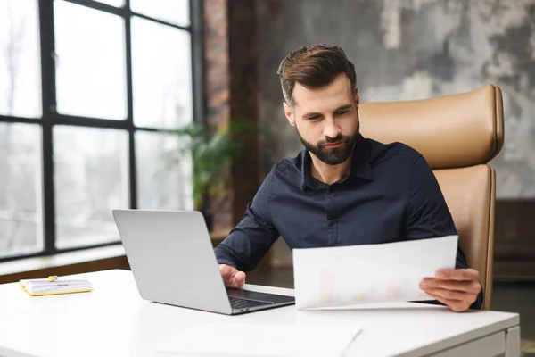 Orolig kaukasisk man tittar igenom korrespondens. Killen som höll dokument i handen, fick bra med posten. Koncentrerad man sitter vid skrivbordet med en bärbar dator på kontoret — Stockfoto