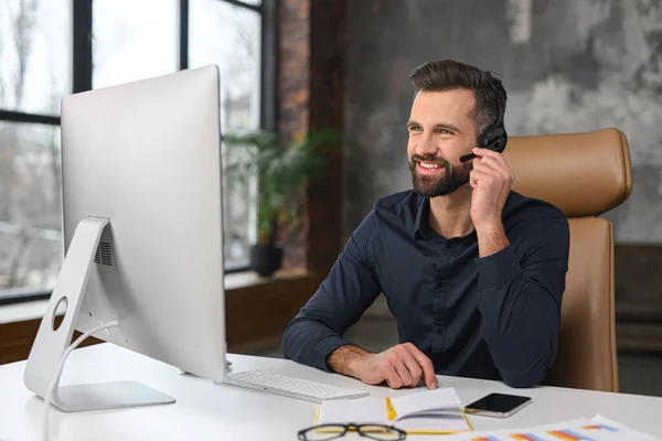 Office employees concept. Focused caucasian man wearing wireless headset is using laptop for online video conversation, talking with customers or coworkers — Zdjęcie stockowe