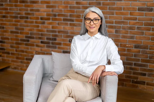 Portrét hrdé a profesionální šedovlasé zralý obchodnice ve stylových brýlích a ležérní oblečení sedí v podkrovní kanceláři. Senior female employee looking to the camera — Stock fotografie