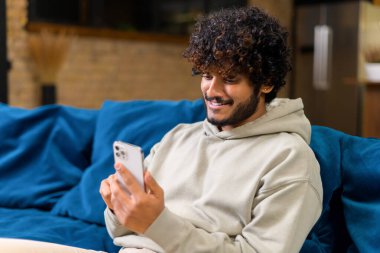 Hintli adam akıllı telefon kullanıyor. Evdeki kanepede oturuyor.
