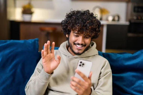 Jeune Indien excité portant un sweat à capuche assis sur le canapé et utilisant un smartphone — Photo