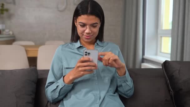 Calma giovane donna in abbigliamento casual seduta sul divano in accogliente soggiorno e tenendo smartphone — Video Stock