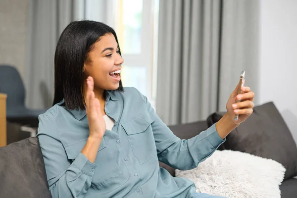 Happy young multiracial teen girl sitting on sofa at home, holding phone, looking at screen and waving hand while video calling distance with friend online. Video chat application — Stockfoto