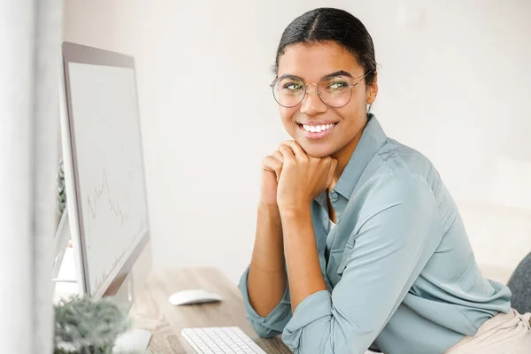 Portrait of young African American woman sitting at the desk with computer, typing email, messaging friend, chatting online with a pleasant smile. Distance work — стокове фото