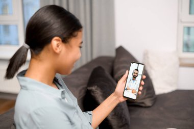 Uzaktaki video bağlantısı. Doktorla görüntülü konuşma yapmak için akıllı telefon kullanan genç bir kadın. Gülümseyen kadın evdeki kanepede otururken internette sohbet ediyor.