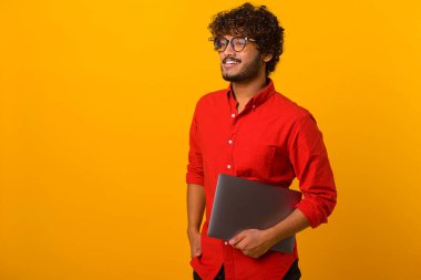 Dizüstü bilgisayarı elinde tutan ve bir yerlere bakan neşeli, kıvırcık latin adamın portre görüntüsünü yükseltin. Kapalı stüdyo çekimleri turuncu renkte.