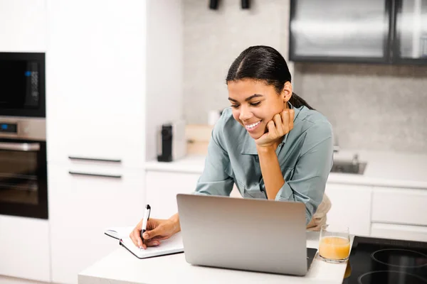 Positive smart young mixed-race female student smiling while standing at the table with laptop in the kitchen, studying from home and taking notes to the notebook, watching an online webinar — Stockfoto