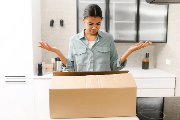 Disappointed young African American girl standing at the kitchen with carton box and feeling upset and confused with the wrong delivery from an online store — 图库照片