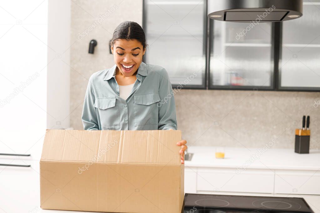 Multiracial woman unpacking parcel cardboard box with online order. Satisfied biracial young woman unpacking parcel box while standing at the kitchen at home