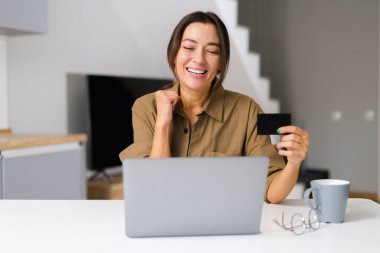 Excited Asian woman shopping online sitting with credit card at home office