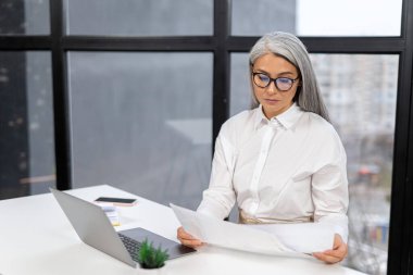 Happy mature businesswoman accountant in glasses sitting at the table with laptop, taking notes, holding papers, contract studying or working online