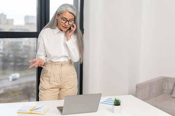 Senior mature gray-haired female professional leader in glasses standing and chatting via smartphone near the office desk — Stok fotoğraf