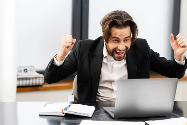 Crazy excited lucky young bearded businessman sitting at the desk with laptop — Stock Photo, Image