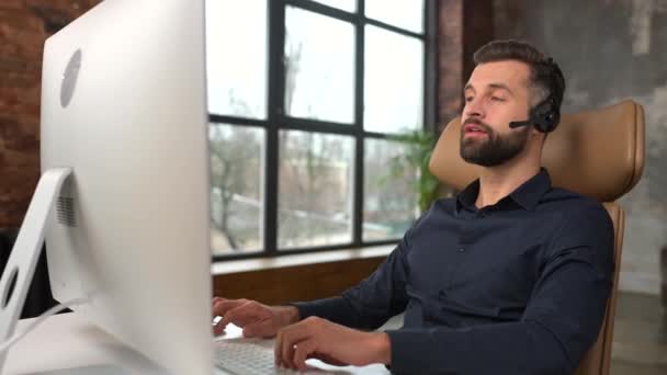 Cheerful guy using handsfree headset and computer to talking online at his workplac — Video Stock