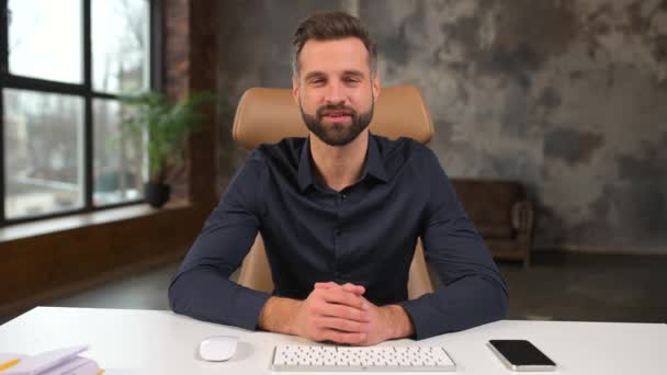 Smiling bearded caucasian businessman sitting at the desk and looking at the camera while waving — Stock video