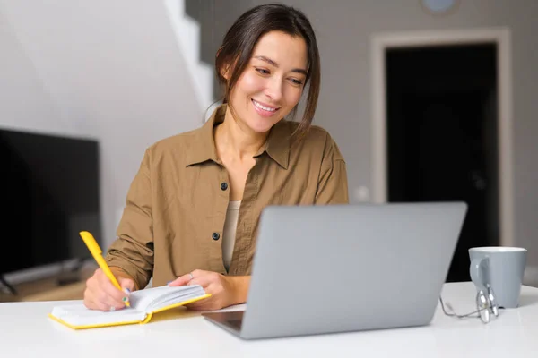 Smiling kind young woman works from home. She sitting at the kitchen table In front of the laptop and notebook — Stockfoto