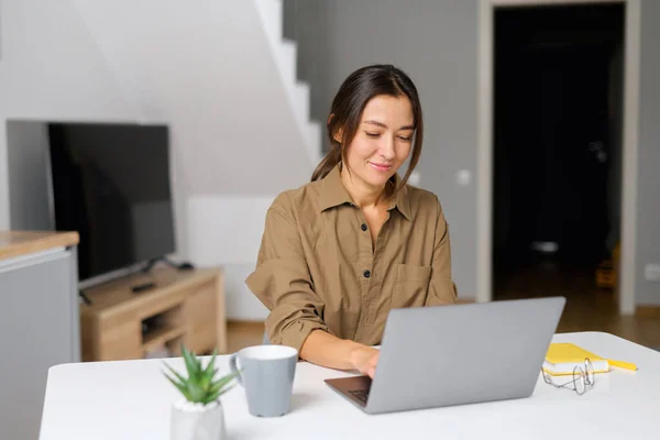 Sonriente joven alegre asiática freelancer disfrutando del café de la mañana mientras trabaja en el portátil, mirando a la pantalla y sentado en el escritorio moderno —  Fotos de Stock