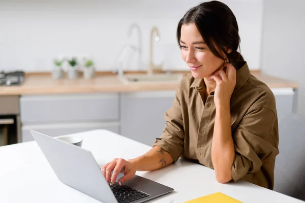 Cheerful asian girl using laptop for remote work or home leisure while sitting in the table at home. Side view of the nice girl looking at the screen — Stok fotoğraf