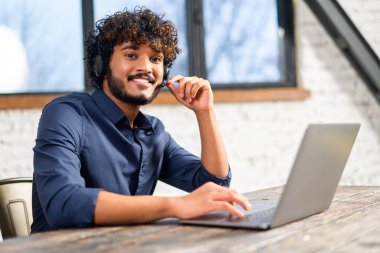 Indian mixed race man wearing wireless headset in smart casual shirt using laptop while sitting at the desk. Inspired Hindu male employee in headphones