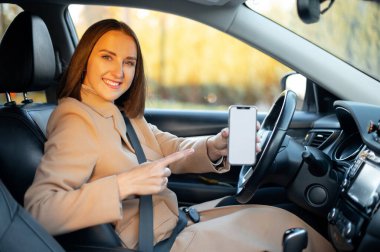 Smiling woman sits in the car and showing smartphone with empty blank screen