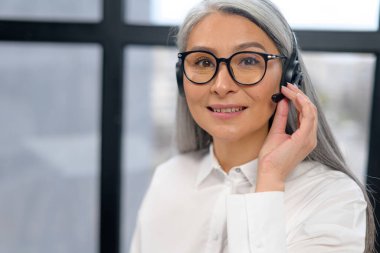 Headshot portrait of a smiling elegant senior lady, wearing headset and glasses looking at the camera. Portrait of a gray-haired customer service representative sitting in the office