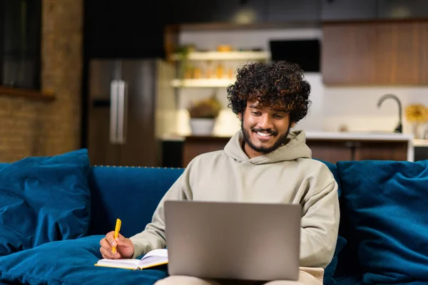 Busy multi-ethnic Hindu freelancer guy using laptop computer for work or studying on the distance from home. Concentrated Indian man watching online webinar and taking notes in notebook — Stock Photo, Image