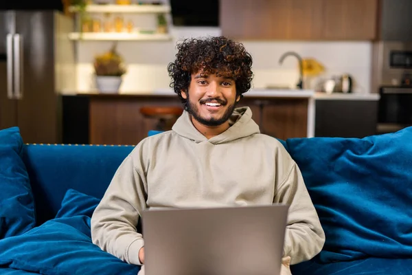 Optimistic confident dark-haired Indian handsome guy using laptop for work. Mixed-race eastern freelancer man sitting at the sofa and looking at camera — 스톡 사진
