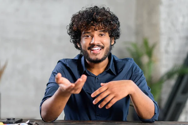 Portrait of happy young Indian guy sitting at desk and gesturing. Smiling curly ethnic man in casual shirt involved virtual meeting. Male student taking a part in educational conference — 图库照片