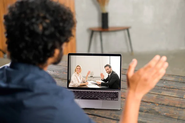 Waist up view of the young man in blue shirt waving with hand and saying hello while chatting via laptop with male and female colleagues — Fotografia de Stock