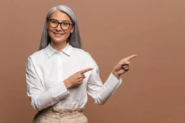 Wow, look at ads here. Portrait of asian elderly woman pointing aside, showing blank copy space for idea presentation, commercial text. Indoor studio shot isolated on beige — ストック写真