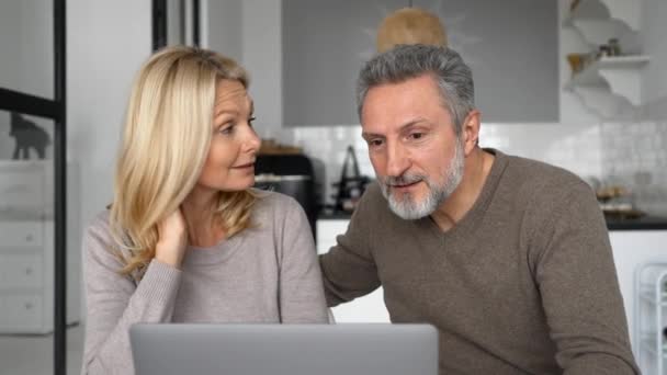 Alegre pareja de mediana edad con un ordenador portátil sentado en el escritorio en la cocina moderna en casa — Vídeos de Stock