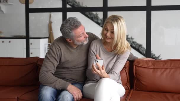 Hilarious mature couple sitting on the sofa and holding smartphone — Vídeo de Stock