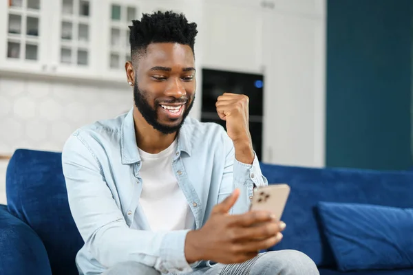 Spännande svart multiracial man sitter på soffan tittar på telefonen och ler, förvånad känslomässig lycklig man, glädjande över bra budskap, nyheter — Stockfoto