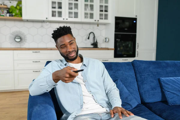 Glad ung multiracial man som håller TV fjärrkontroll medan du sitter på en soffa hemma och koppla av på helgen — Stockfoto