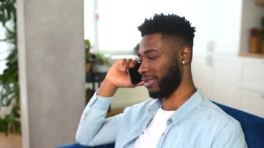 Mutlu yakışıklı Afro-Amerikalı adam evdeki kanepede oturup eğlenceli telefon konuşmaları yapıyor.