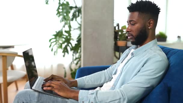 Sorrindo afro-americano freelancer cara está usando computador portátil sentado no sofá em casa — Vídeo de Stock