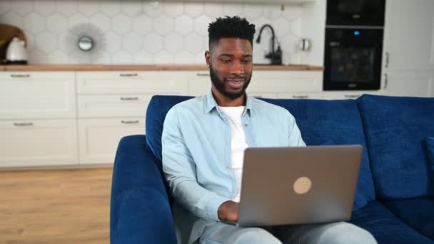 Homem alegre vestindo roupas casuais sentado no sofá e usando laptop em casa — Vídeo de Stock