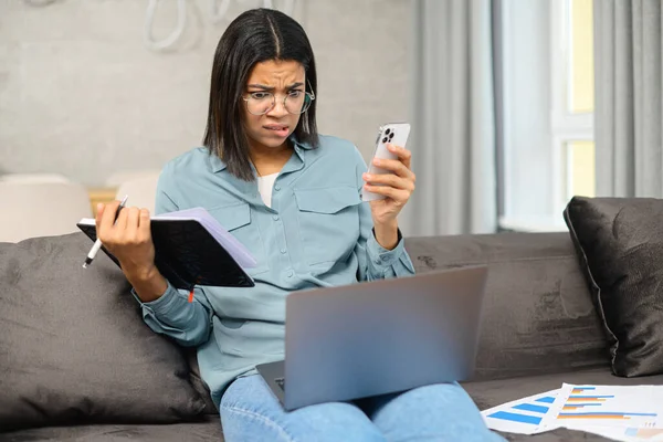 Konzentrierte junge afroamerikanische Studentin mit Brille sitzt auf der Couch, Laptop auf dem Schoß, lernt von zu Hause aus mit aufmerksamem Gesicht — Stockfoto